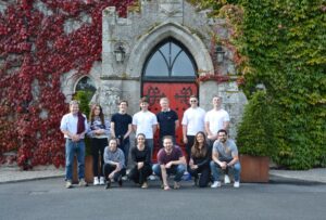 Ronan Group Real Estate team at Barretstown Castle - 12 staff members group photo in front of historic ivy-covered stone building with Gothic arched entrance and red doors. Dublin property development company community involvement visit to children's charity castle